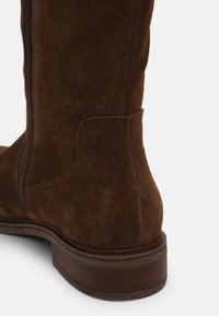 Bota alta hasta la rodilla de ante marrón con cremallera lateral, textura suave, suela de cuero y diseño de punta redonda. Acentuada con una costura cosida.