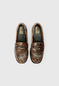 Mocassins de pele de cobra castanha com ponta arredondada, apresentando uma faixa de sela preta e um detalhe em logo dourado na parte superior. Forro interior suave.