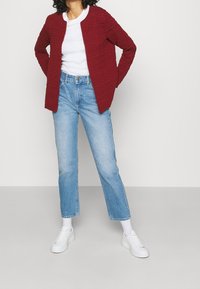 Chaqueta roja texturizada de corte recto, llevada sobre una camisa blanca, combinada con pantalones vaqueros de un azul claro y zapatillas blancas.