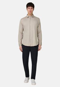 Beiges langärmliges Hemd mit Knöpfen und Kragen, kombiniert mit einer schwarzen Hose und weißen Turnschuhen, vor einem einfarbigen Hintergrund stehend.