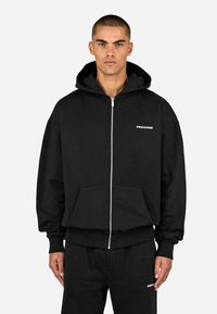 Sweatshirt preta com fecho de correr, feita de algodão, com bolso frontal, apresentando um logótipo branco simples no lado esquerdo do peito e punhos e bainha canelados.