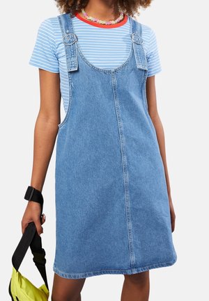 Femme portant une robe salopette en denim bleu par-dessus un t-shirt rayé à manches courtes, tenant un sac jaune avec une sangle noire dans une main.