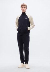 Αθλητική φόρμα σε ναυτικό μπλε και μπεζ, κατασκευασμένη από μαλακό ύφασμα με ζακέτα με φερμουάρ, δύο πλαϊνές τσέπες και αντίστοιχο παντελόνι· διακοσμημένη με λευκές λογότυπες λεπτομέρειες.