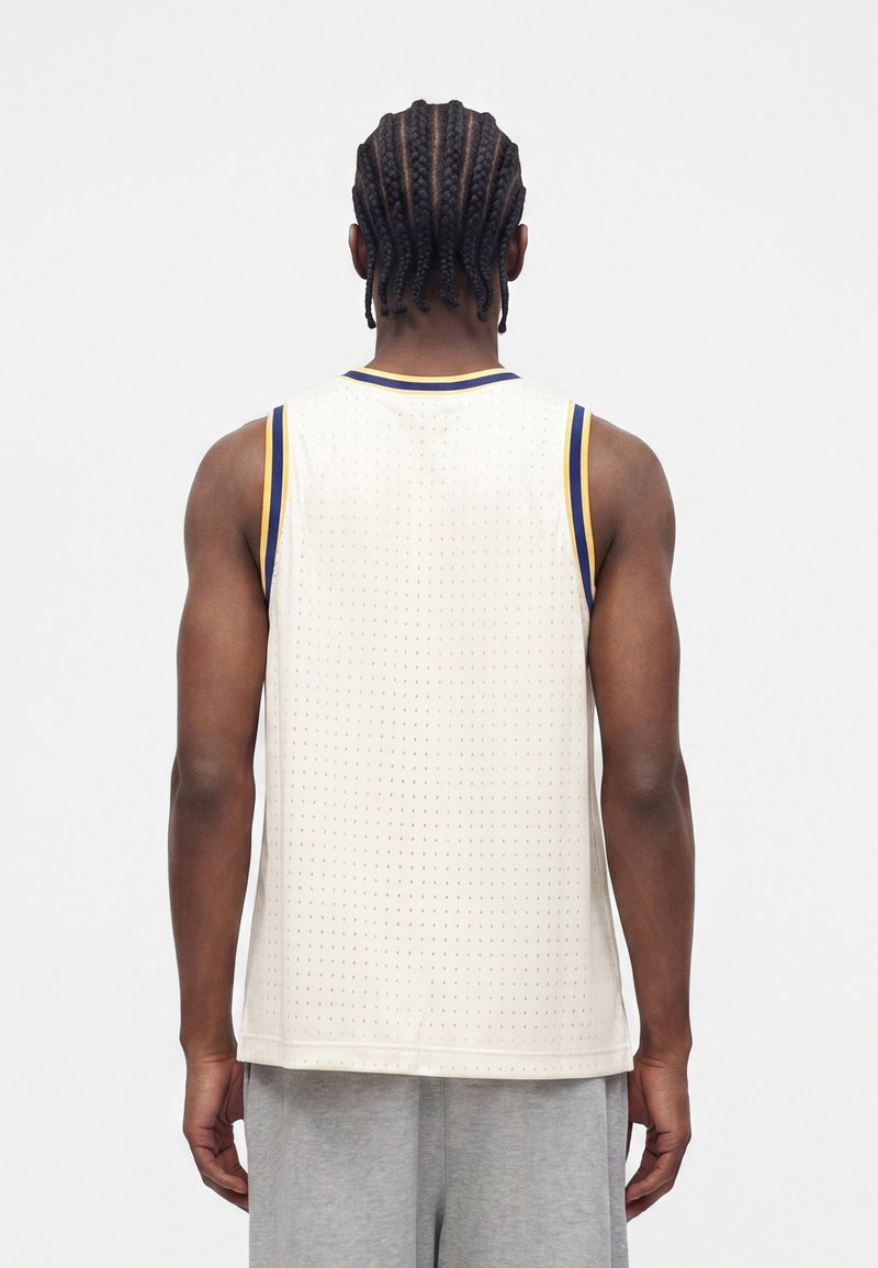 Person with braided hair stands facing away wearing a sleeveless white sports jersey with blue and yellow trim and gray pants.