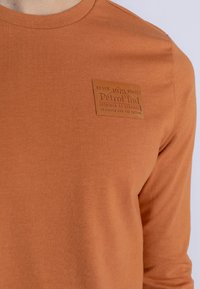 Långärmad t-shirt i rostfärg med mjukt tyg som har en strukturerad brun läderlapp med varumärke på bröstet.