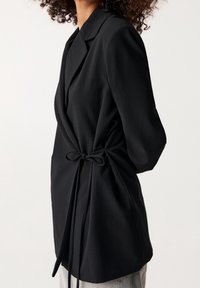 Veste noire portefeuille avec taille nouée, en tissu lisse, col à revers et manches longues, conçue pour une silhouette élégante.