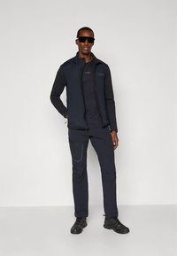 Svart multifunktionell väst över en svart långärmad tröja, kombinerad med mörka byxor och svarta skor. Har subtil branding och en stilren design.