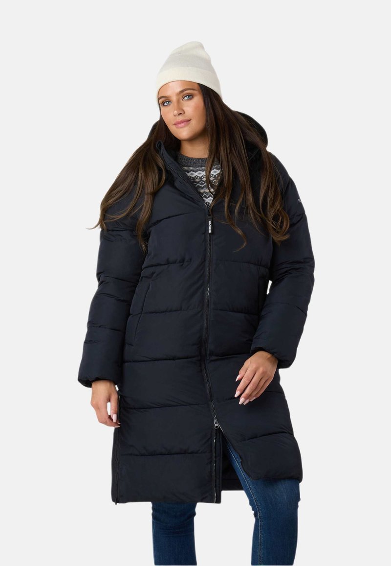 Sort puffjakke med lynlås foran og hætte, med quiltet design, lange ærmer og sidelommer. Båret sammen med en grå mønstret sweater.