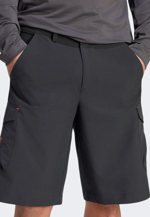 Mann trägt schwarze Cargoshorts und ein langärmliges graues Hemd, die Hände in den Hosentaschen, Bild auf Taille und Oberschenkel beschnitten.