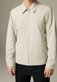 Lichtbeige jas met rits, een puntige kraag, lange mouwen en een gladde textuur. Gecombineerd met donkere spijkerbroek.