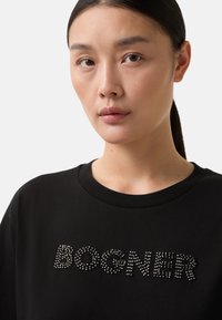 Ung kvinde med mørkt hår samlet i en hestehale, iført en sort skjorte med naglede bogstaver, der staver "BOGNER" på brystet.