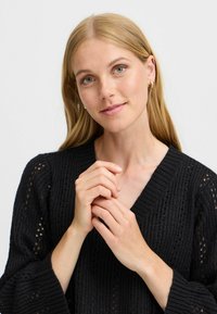 Un pull en tricot noir avec une encolure en V, des manches amples et un design perforé. Un modèle avec de longs cheveux et une expression neutre tient ses mains ensemble.