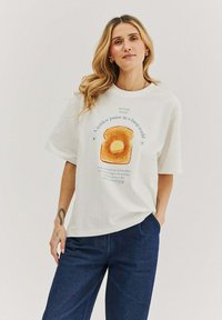Bílé bavlněné tričko s grafickou potiskem toastu v oranžových a hnědých odstínech, s nápisy nahoře a dole, v uvolněném střihu, doplněné tmavě modrými kalhotami.