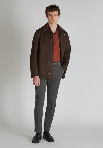 Giacca di cotone marrone con colletto, indossata sopra un maglione a maglia color ruggine, abbinata a pantaloni grigi slim e scarpe nere.