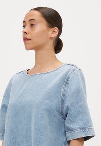 Camisa de denim azul clara com decote redondo, mangas curtas e costuras visíveis ao longo das costuras e bordas.