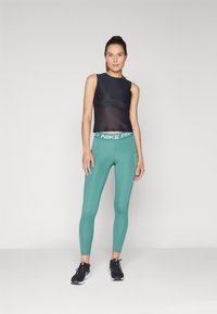 Črn brezrokavnik z gladkim zaključkom v kombinaciji z zelenimi Nike ProLeggingami, ki imajo bel pas in stranske žepe. Črne atletske čevlje.
