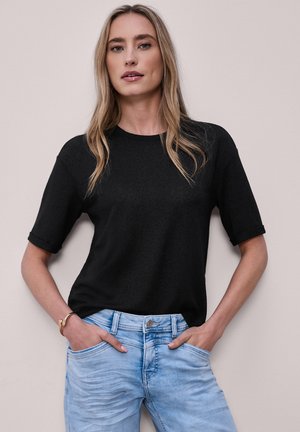 Kobieta z długimi blond włosami, ubrana w czarną koszulkę z krótkim rękawem i jasnoniebieskie dżinsy, stojąca z rękami w kieszeniach na jednolitym tle.