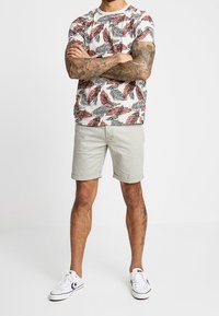 Camiseta blanca de manga corta con un patrón de hojas en rosa y verde, combinada con pantalones cortos grises claros y zapatillas blancas.