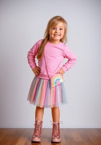 Rosa Sweatshirt mit langen Ärmeln, geschichteter pastellfarbener T�üllrock, glänzende rosa Stiefel und eine Handtasche mit Regenbogenmuster. Steht auf einem Holzboden.