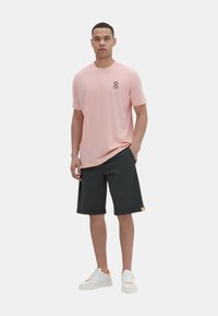T-shirt rosa chiaro a maniche corte con logo piccolo, abbinato a pantaloni corti grigi scuri di lunghezza al ginocchio. Sneakers bianche con dettagli arancioni completano l'outfit.