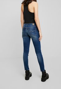 Jeans de mezclilla azul ajustados con bolsillos traseros; etiqueta en la cinturilla; combinados con una camiseta sin mangas negra y botas negras hasta el tobillo. Textura suave.