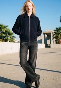 Tânără stă afară cu mâinile în buzunare, purtând o geacă neagră cu fermoar, blugi închiși la culoare și pantofi negri sub un cer albastru clar.