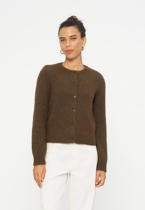 Femme aux cheveux bouclés en chignon, portant des boucles d'oreilles anneaux dorées, un cardigan marron boutonné et un pantalon blanc, debout devant un fond blanc uni.