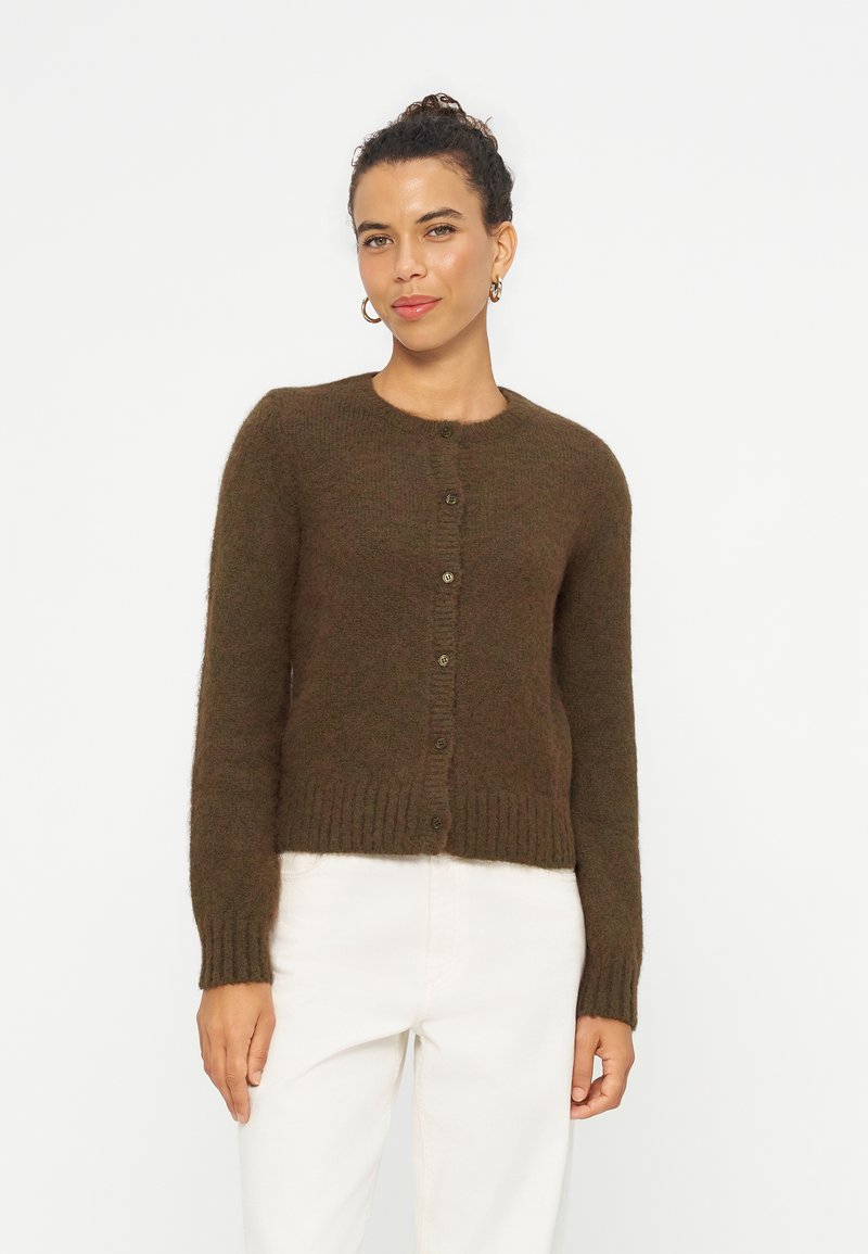 Femme aux cheveux bouclés en chignon, portant des boucles d'oreilles anneaux dorées, un cardigan marron boutonné et un pantalon blanc, debout devant un fond blanc uni.