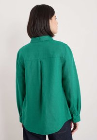 Camicia di lino verde con colletto, maniche lunghe e orlo curvo. Presenta una piega sul retro e polsini abbottonati.