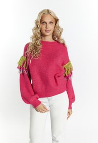 Fukszia kötött pulóver, oversized ujjal és zöld, rózsaszín, illetve szürke bojtokkal a vállán. Világos színű farmernadrággal párosítva.