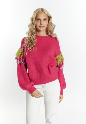 Pull en tricot fuchsia avec des manches surdimensionnées et des accents de franges vertes, roses et grises sur les épaules. Associé à un jean de couleur claire.