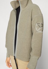 Giacca in fleece colore oliva chiaro, con colletto alto, zip completa e superficie texture. Logo accentato sulla manica sinistra; polsini elasticizzati.