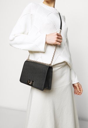 A person wearing a textured white sweater holds a black handbag with a chain strap, standing against a light background.