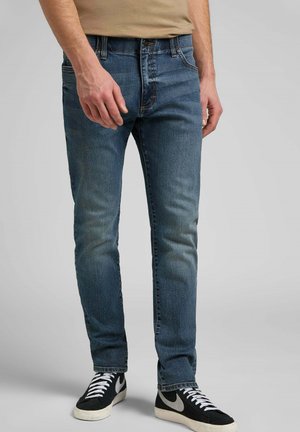 Jeans in denim blu slim fit con lieve sbiaditura, design a cinque tasche e cuciture a contrasto. Indossati con sneakers nere caratterizzate da accenti bianchi.
