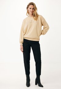 Sudadera beige con letras brillantes "MEXX", combinada con pantalones negros ajustados y botas de tacón negras. Textura de tela suave.