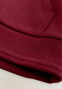 Fondo di felpa bordeaux con texture a costine. Presenta una composizione di tessuto morbido e una cucitura doppia lungo il bordo.