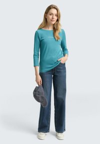 Türkises Langarmoberteil mit Rundhalsausschnitt und weißem Text; kombiniert mit weitem blauen Jeans. Das Model hält eine graue Kappe. Lässiges Outfit.