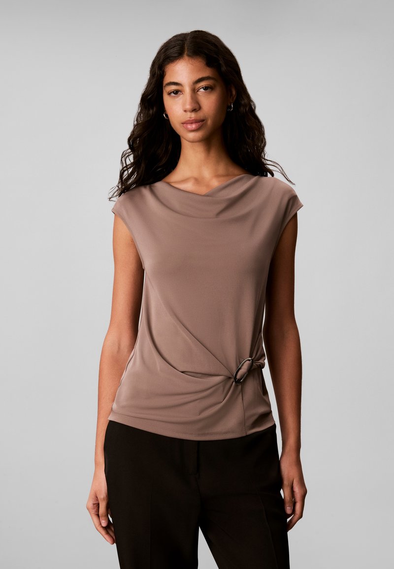 Femme aux longs cheveux foncés portant un haut taupe sans manches avec un col drapé et un détail anneau sur le côté, associée à un pantalon noir.
