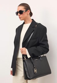 Fekete bőrtáska krokodilbőr mintázattal, arany kiegészítőkkel és díszes zárral. A modellen fekete napszemüveg és fekete zakó található.