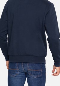 Felpa blu navy con orlo e polsini a coste, caratterizzata da una texture liscia. Abbinata a jeans blu con tasche posteriori visibili.