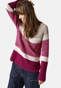 Stickad tröja med horisontella ränder i mörkrosa, ljusrosa och krämfärgad. Har en bred halsringning och ribbade ärmslut, kombinerad med mörka jeans.