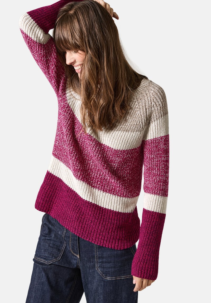Stickad tröja med horisontella ränder i mörkrosa, ljusrosa och krämfärgad. Har en bred halsringning och ribbade ärmslut, kombinerad med mörka jeans.