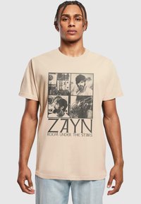 Krémové bavlněné tričko s grafickým designem obsahujícím čtyři monochromatické obrázky a text "ZAYN ROOM UNDER THE STAIRS" napsaný tučným písmem.