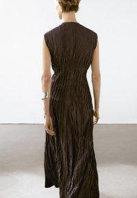 Long, sleeveless dress in dark brown with textured, pleated fabric. Features a high neckline and a flowing silhouette. Silver cuff on arm.