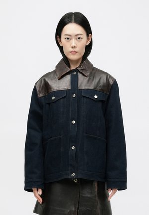 Tamsiai mėlyna striukė su rudos odos pečių akcentais, sidabro spalvos metaliniais mygtukais, dviem priekinėmis kišenėmis ir klasikinio tipo apykakle.