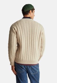 Béžový žebrovaný pletený svetr se středním výstřihem a silným lemem. Doplněný volnými modrými džíny; zadní pohled ukazuje texturovanou látku.