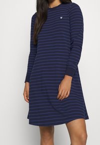 Granatowa sukienka z długim rękawem z poziomymi paskami w jaśniejszym odcieniu niebieskiego. Okrągły dekolt, luźny krój oraz detal z logo na piersi.