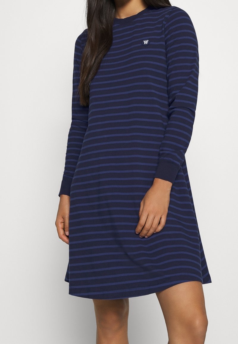 Granatowa sukienka z długim rękawem z poziomymi paskami w jaśniejszym odcieniu niebieskiego. Okrągły dekolt, luźny krój oraz detal z logo na piersi.