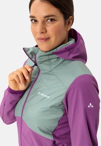 Lila und türkisfarbene isolierte Jacke mit einem Reißverschlusskragen und Logo. Verfügt über eine gesteppten Textur im oberen Bereich und glatte Ärmel.