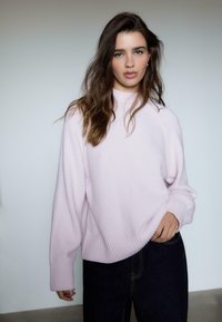 Lyserød strikket sweater med ribbede kanter og manchetter, med høj hals og løs pasform, parret med mørke denim jeans.